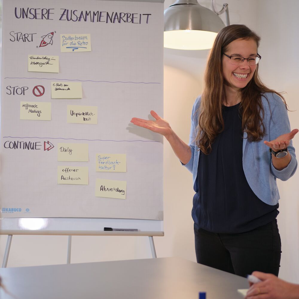 Sarah Werther lacht am Flipchart einer Retrospektive mit Start, Stop, Continue und Haftnotizen